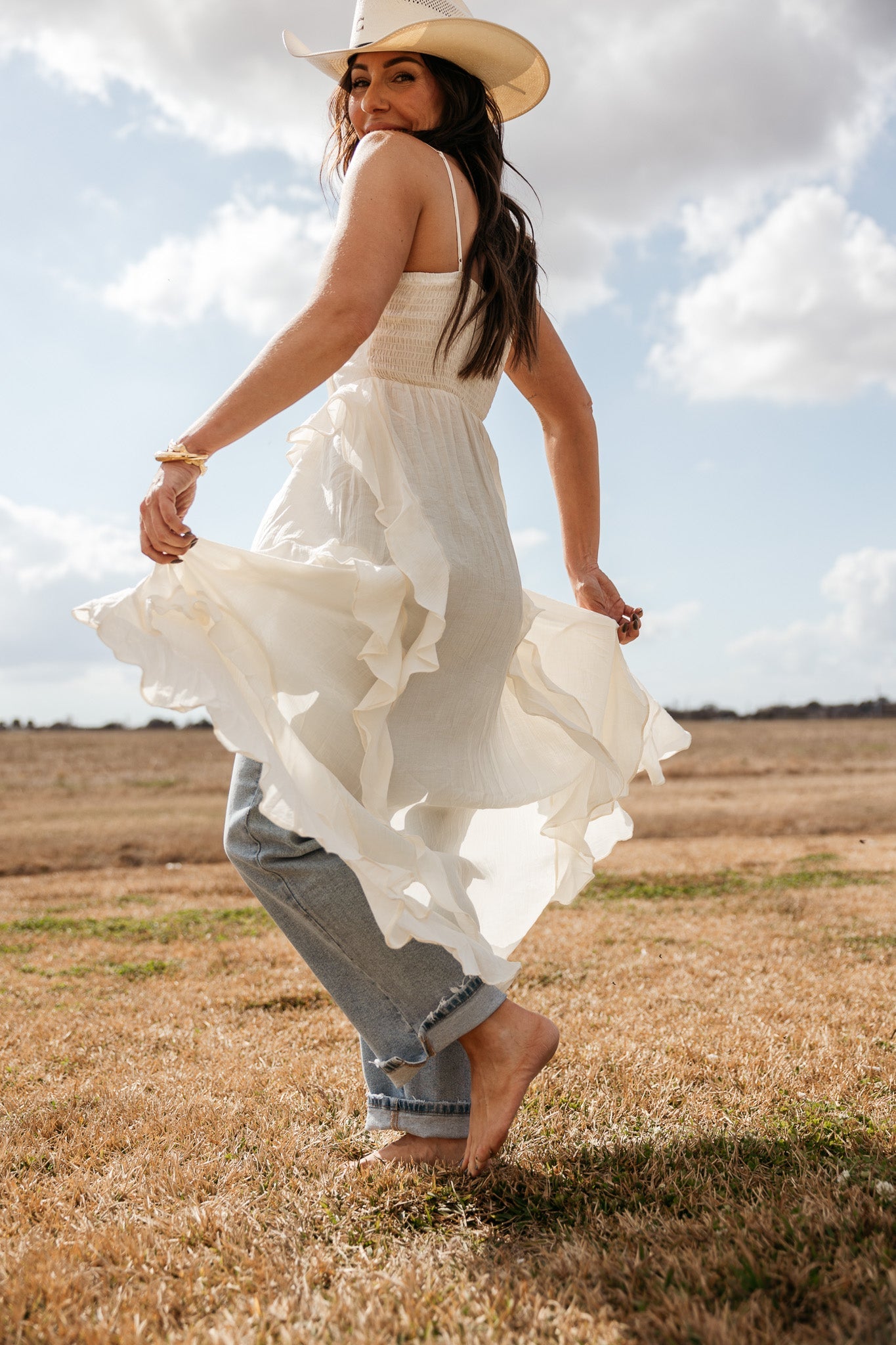 Woman in cowgirl hat, white western boho dress and jeans in a sunny outdoor field