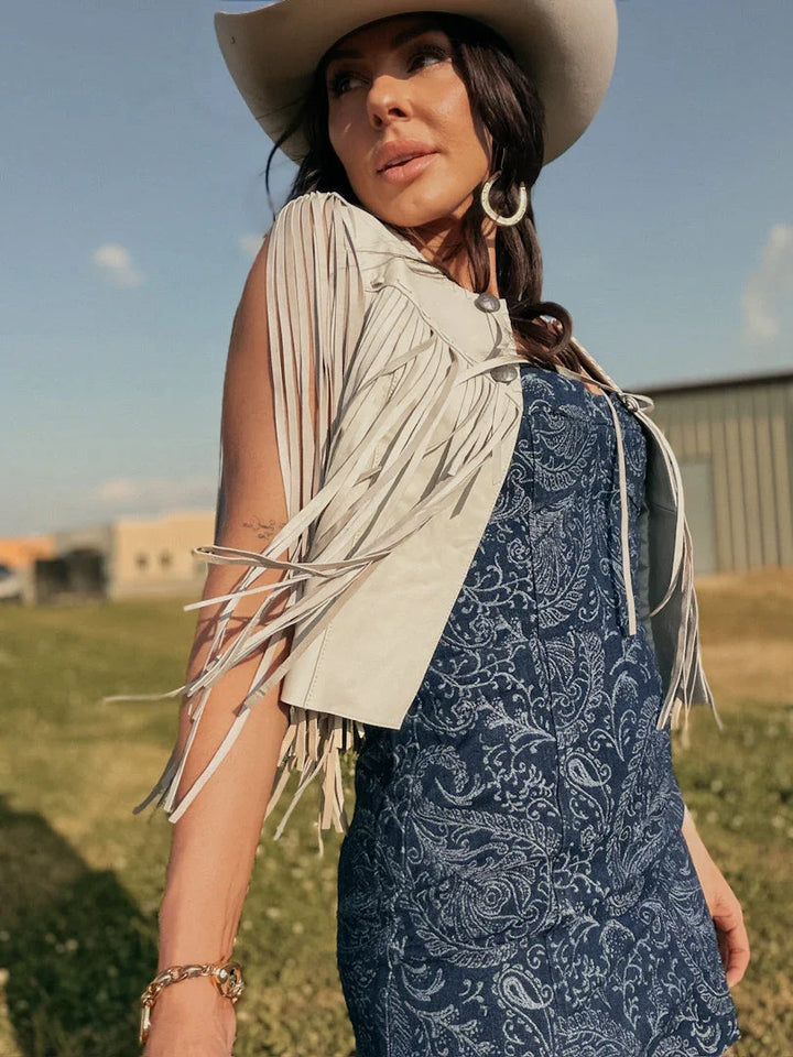 Woman in white fringed vest, blue paisley western dress, and cowgirl hat outdoors