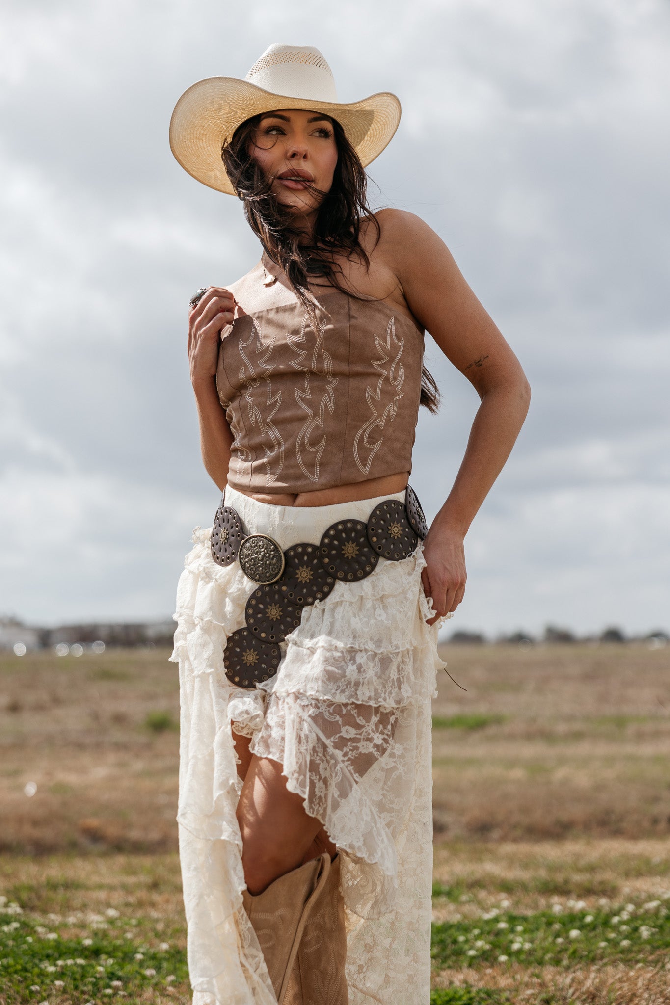Woman in western boho outfit with cowboy hat, brown corset, lace skirt, and concho belt outdoors