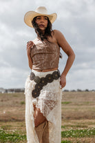 Woman in western boho outfit with cowboy hat, brown corset, lace skirt, and concho belt outdoors