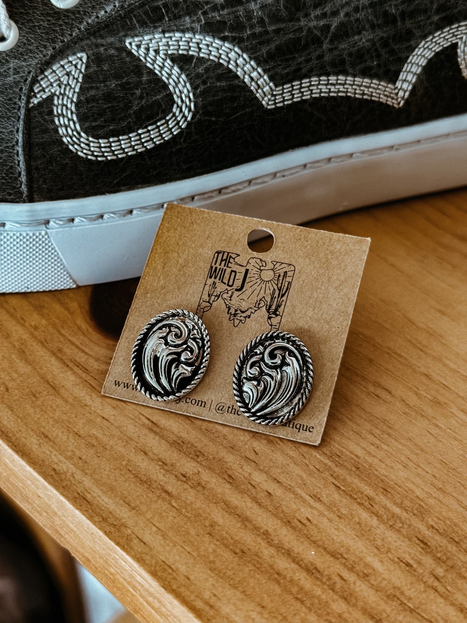 Silver western engraved oval stud earrings on a jewelry card, close to a stitched sneaker.