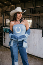 Woman in western boho denim outfit and cowboy hat standing in barn, patchwork jeans, horseshoe top.