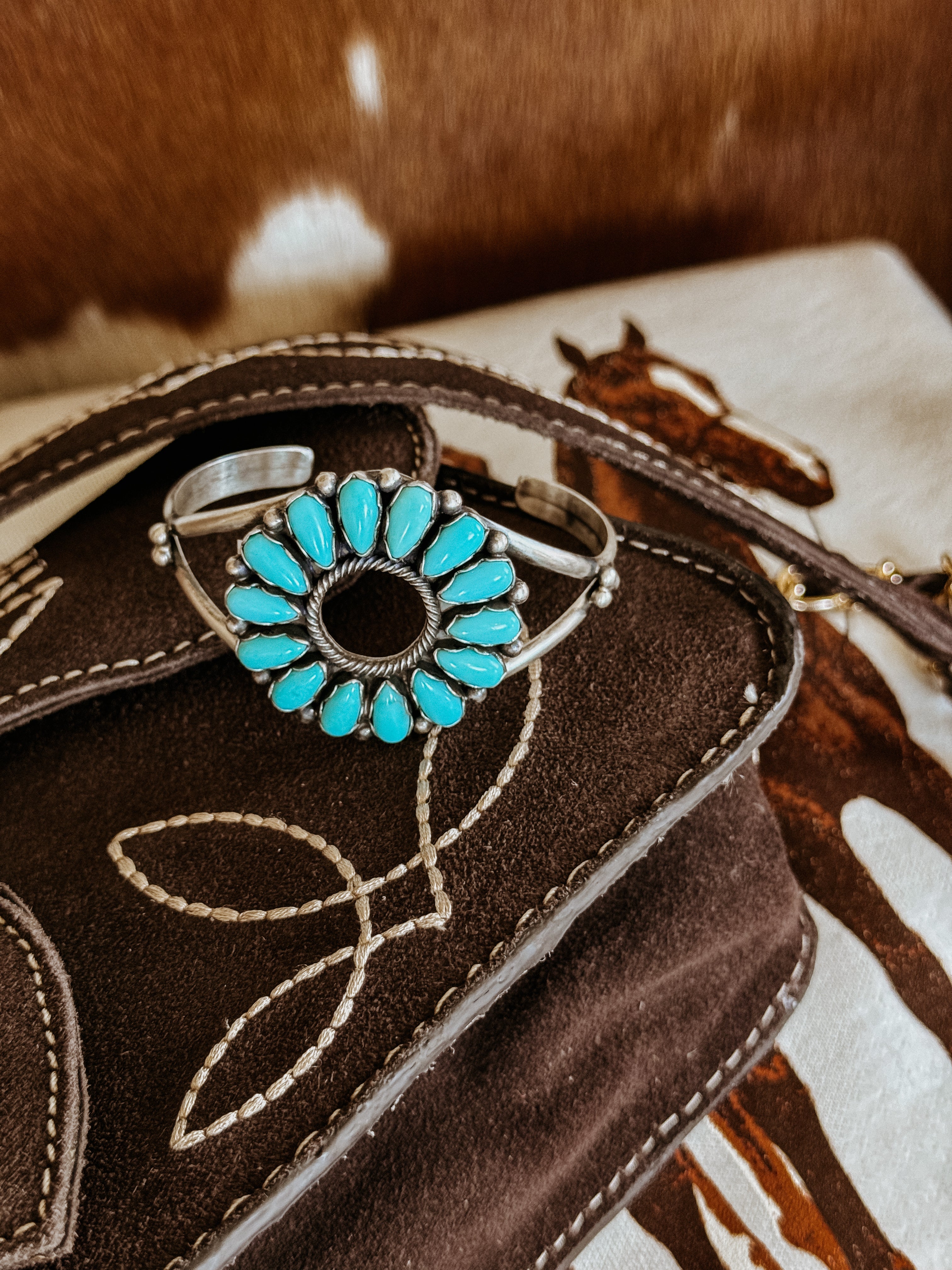 Turquoise stone western cuff bracelet on embroidered suede bag, horse print background.