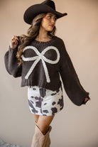 Woman in western cowgirl outfit with black hat, chunky sweater with white bow, cow print skirt, and cowboy boots