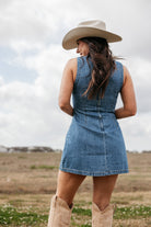 Woman in a denim dress, cowgirl hat, and western boots standing outdoors in a field