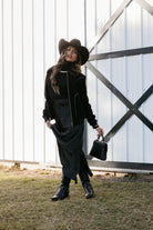 Woman in black cowgirl hat, western jacket, black dress, and boots standing outside barn