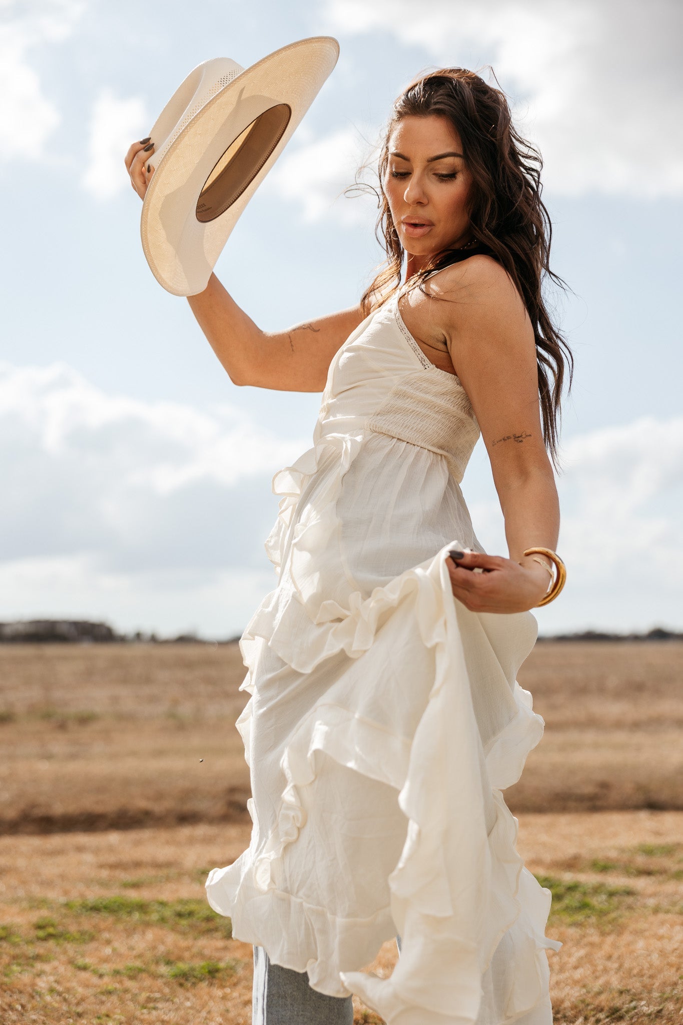 Woman in a flowing white western boho dress holding a hat outdoors in a rustic field