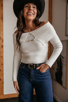 Woman in western off-shoulder white sweater, wide-leg jeans, and black hat smiling indoors