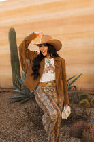 Woman in western boho outfit with cowboy hat, fringe jacket, snakeskin pants, and turquoise jewelry standing outdoors with cactus plants.