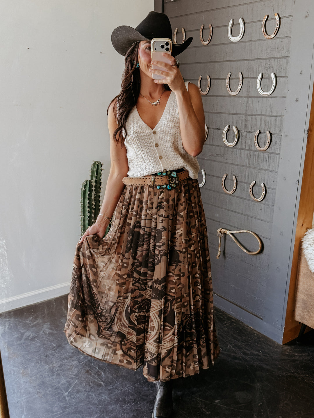 Person wearing a white sleeveless top and patterned skirt with a black hat, standing in front of a wall with decorative horseshoe designs.