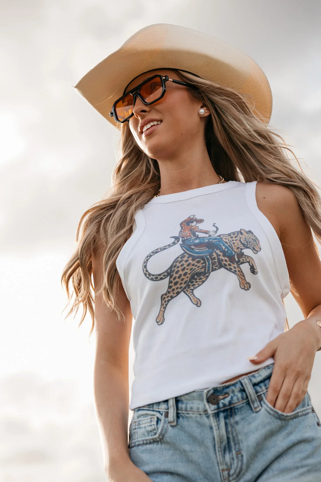 Woman wearing a white tank top with a graphic design, sunglasses, and a wide-brimmed hat against a light background.