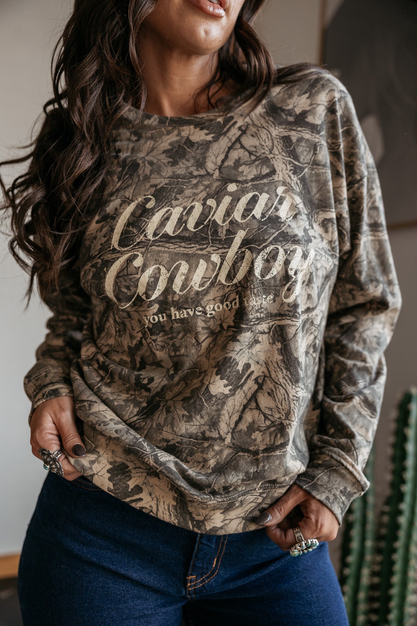 Woman wearing a western boho camo print "Caviar Cowboy" graphic sweatshirt and denim jeans