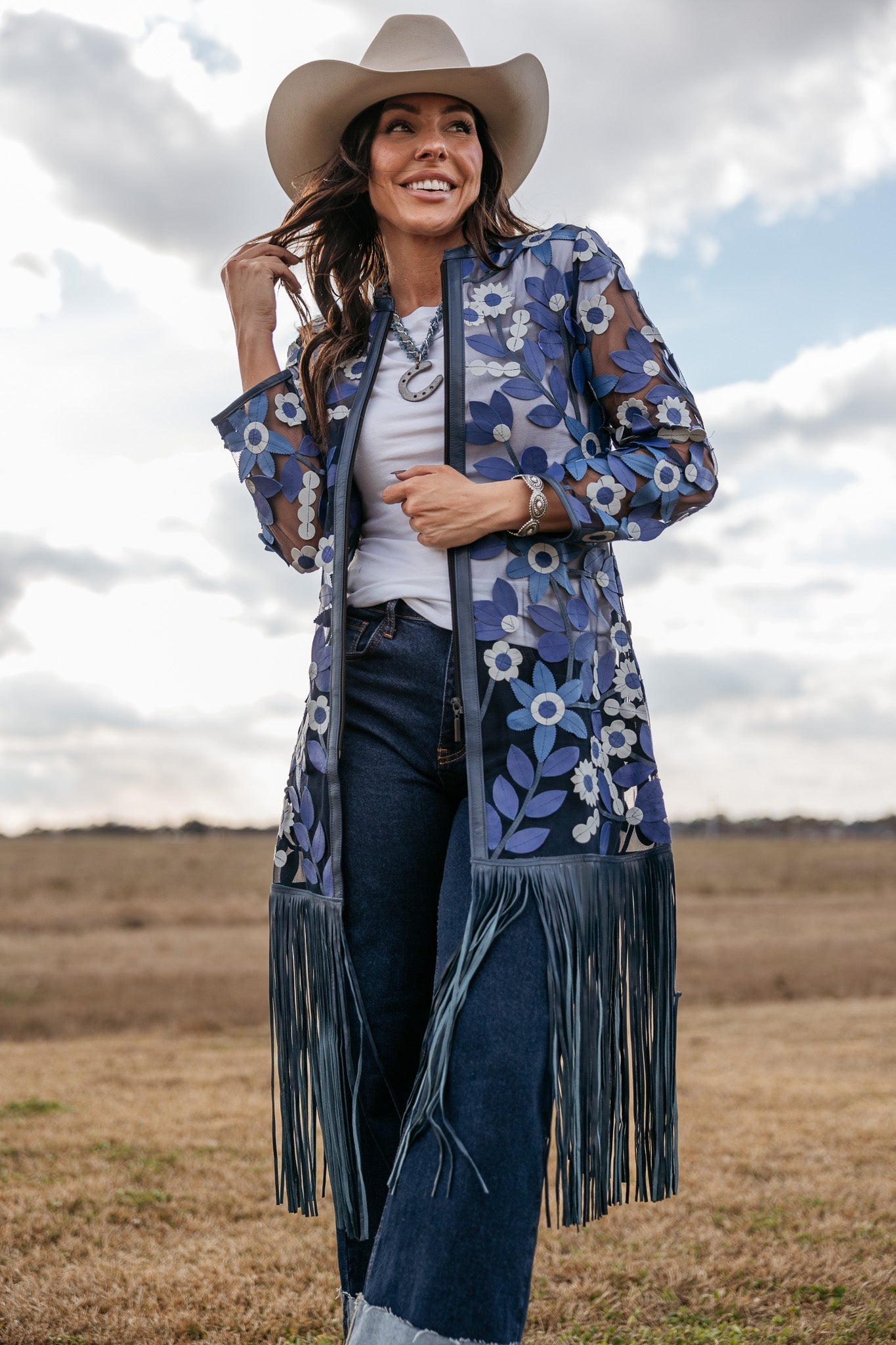 Woman in cowboy hat wearing sheer blue floral fringe duster, white top and jeans outdoors
