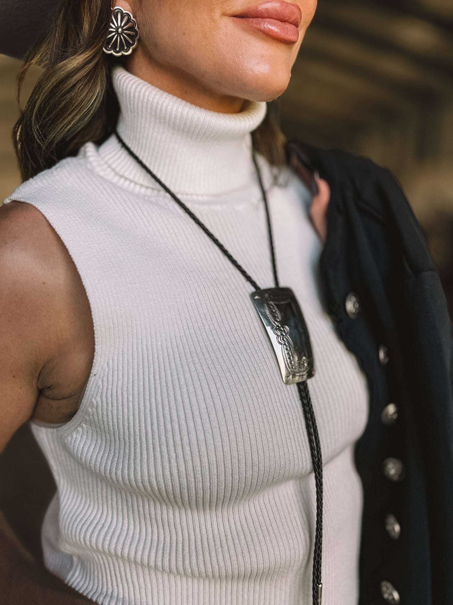 Woman wearing a white ribbed sleeveless turtleneck, western bolo tie, and floral earrings
