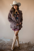 Woman in western cowgirl outfit with wide-brim hat, faux fur jacket, floral dress, and knee-high boots