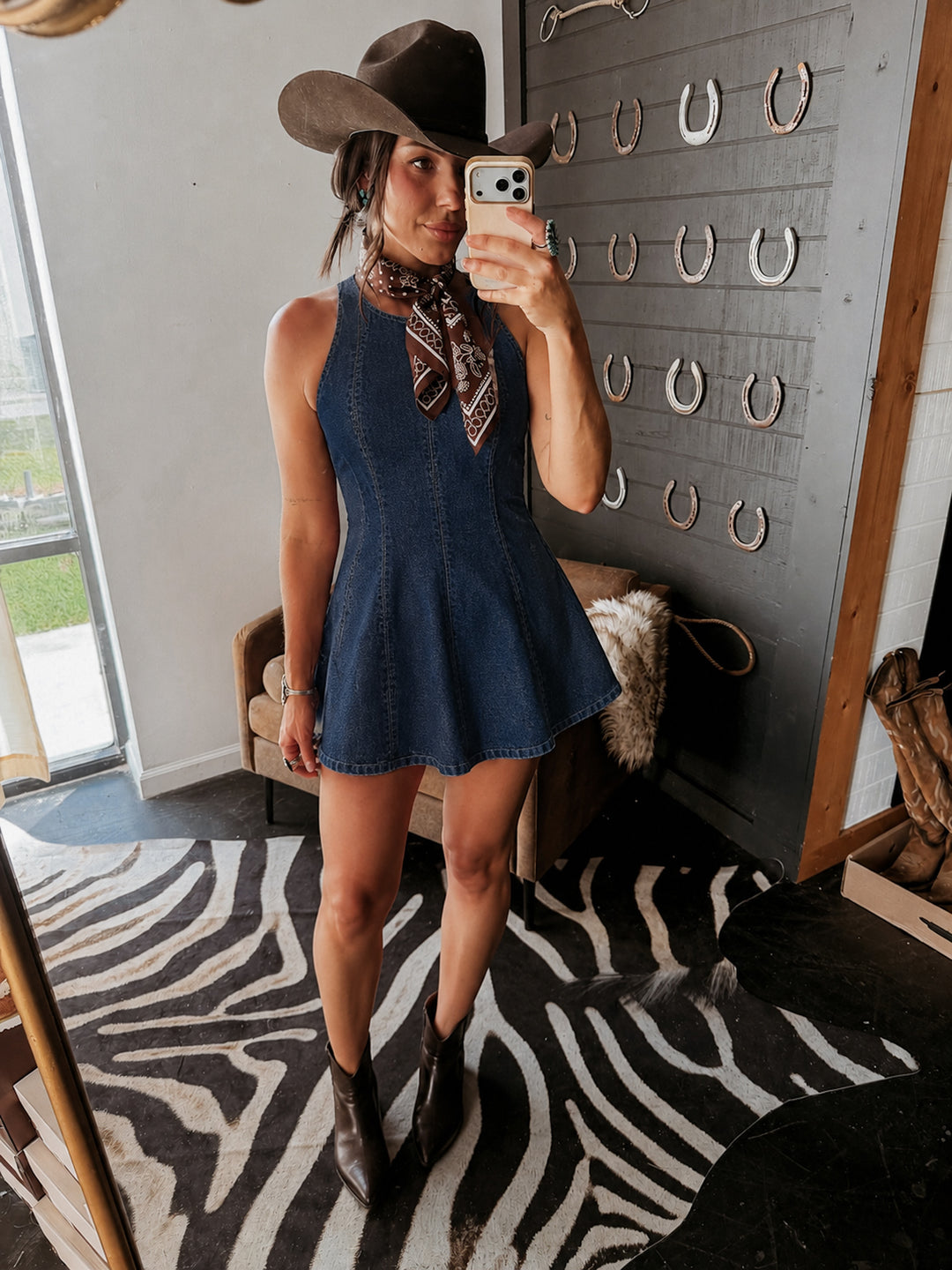 A woman takes a mirror selfie wearing a sleeveless dark blue denim flared romper, a brown cowboy hat, a patterned neck scarf, and brown ankle boots. She is standing in a room with a zebra-print rug, a gray wall decorated with horseshoes, and a window on the left.
