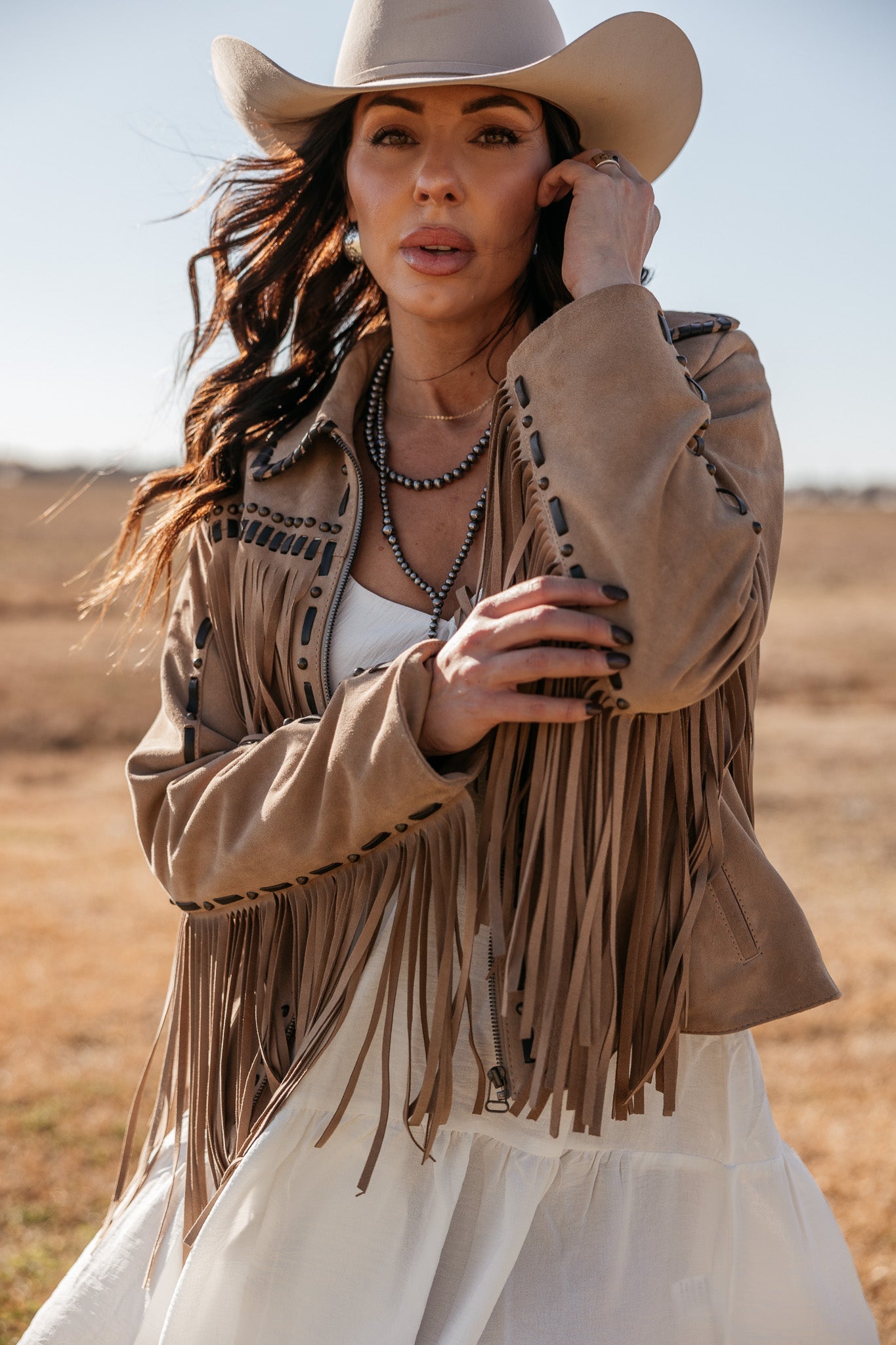 Woman in tan fringe suede jacket, white dress, and cowboy hat in western boho fashion style outdoors