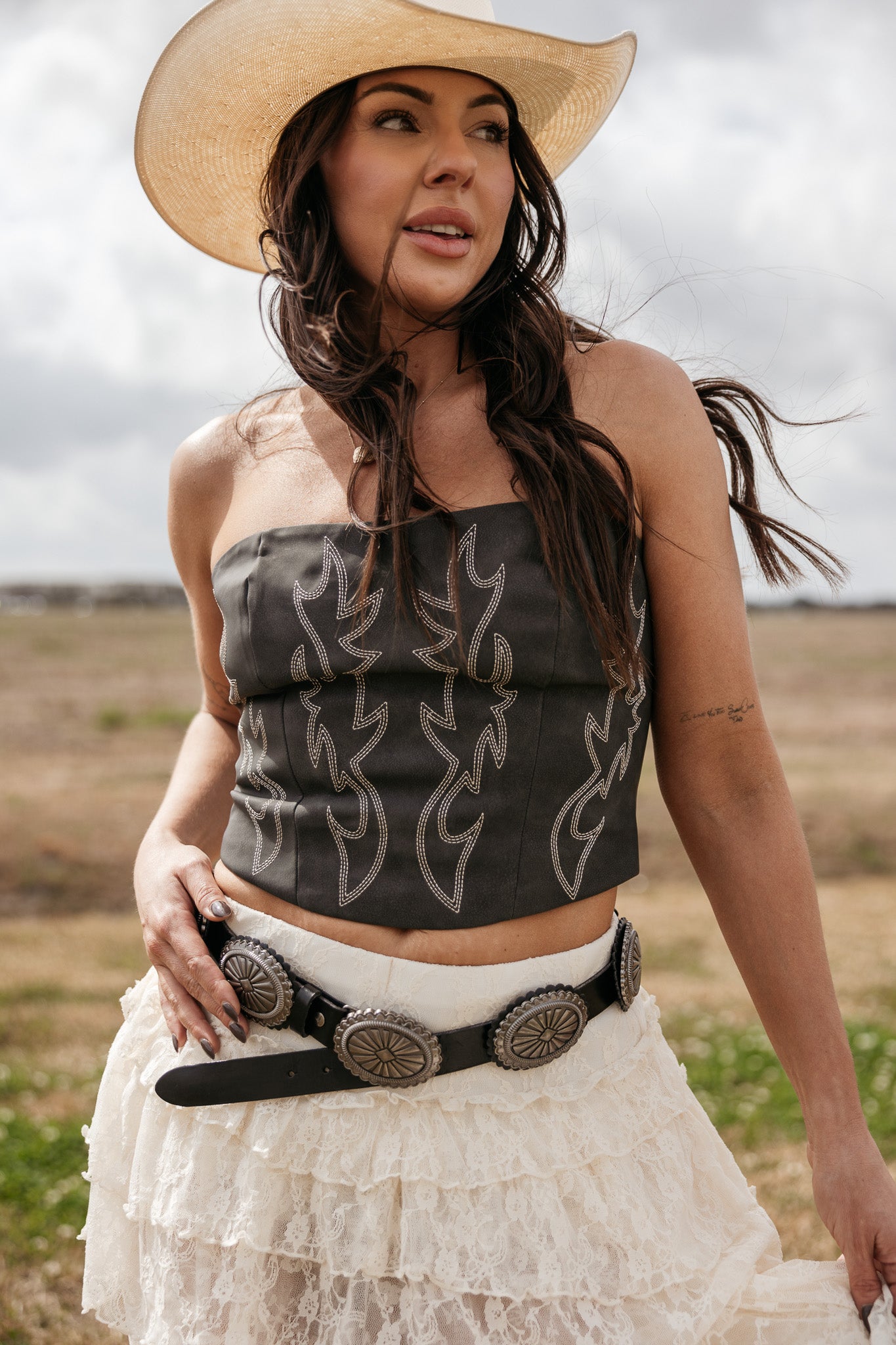 Woman in a western cowgirl outfit with a cowboy hat, black embroidered corset, lace skirt, and concho belt outdoors.