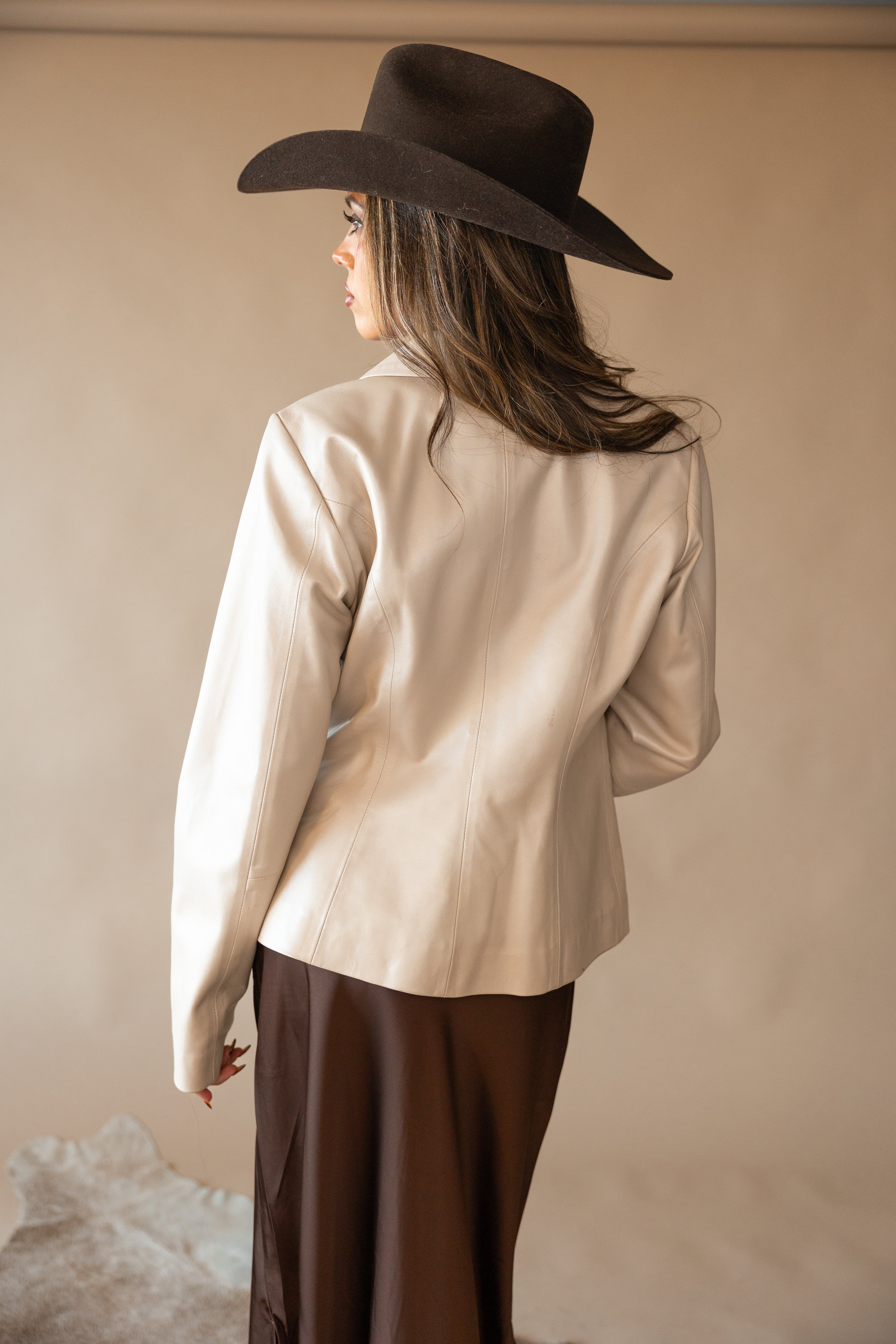 Woman in brown cowgirl hat, cream western jacket, and brown skirt facing away