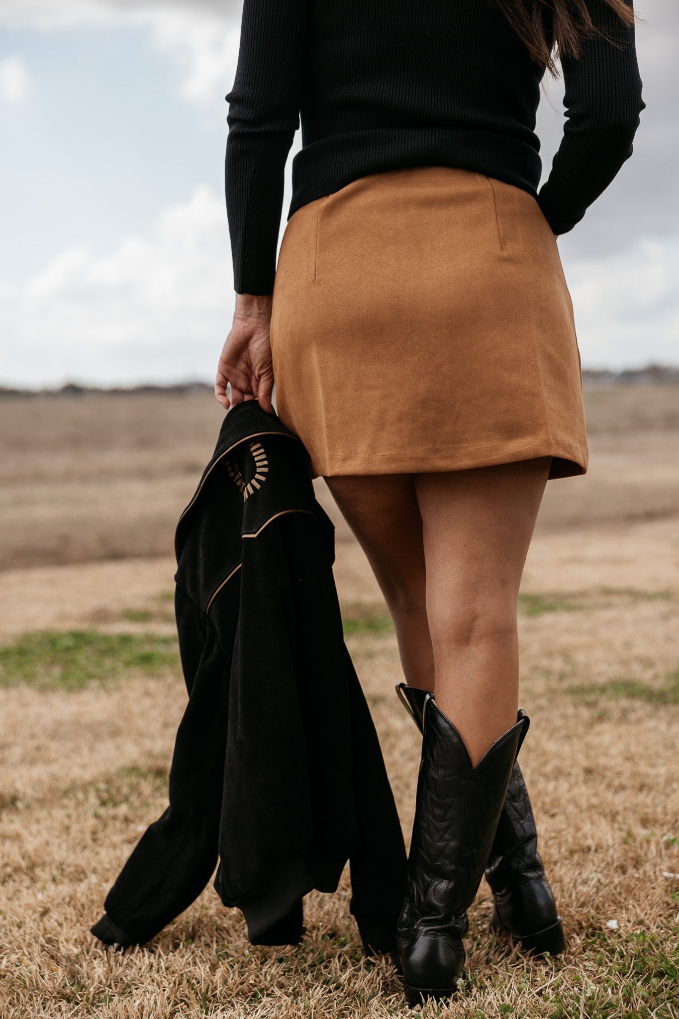 Woman in brown suede western mini skirt and black cowboy boots outdoors, holding a jacket
