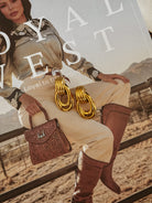 Gold chunky hoop earrings displayed on a western fashion magazine featuring a cowgirl in embroidered blouse, cream pants, tooled leather purse, and brown knee-high boots.