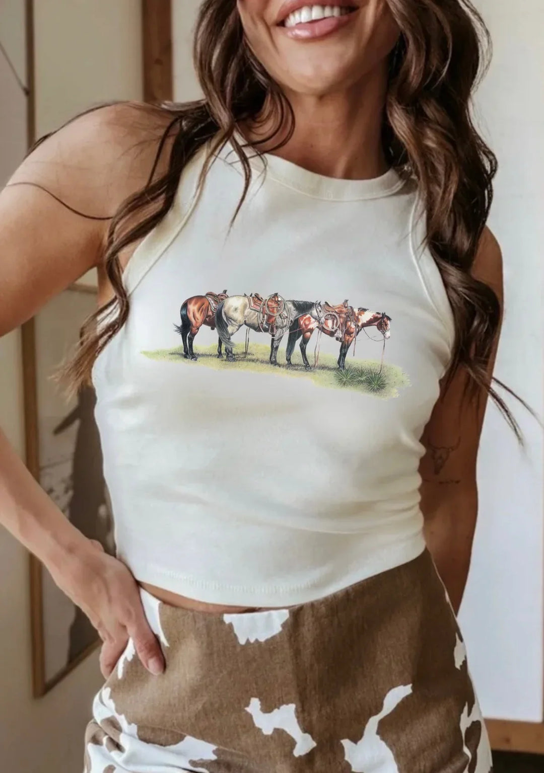 Woman wearing a white tank top with horse design and brown pants.