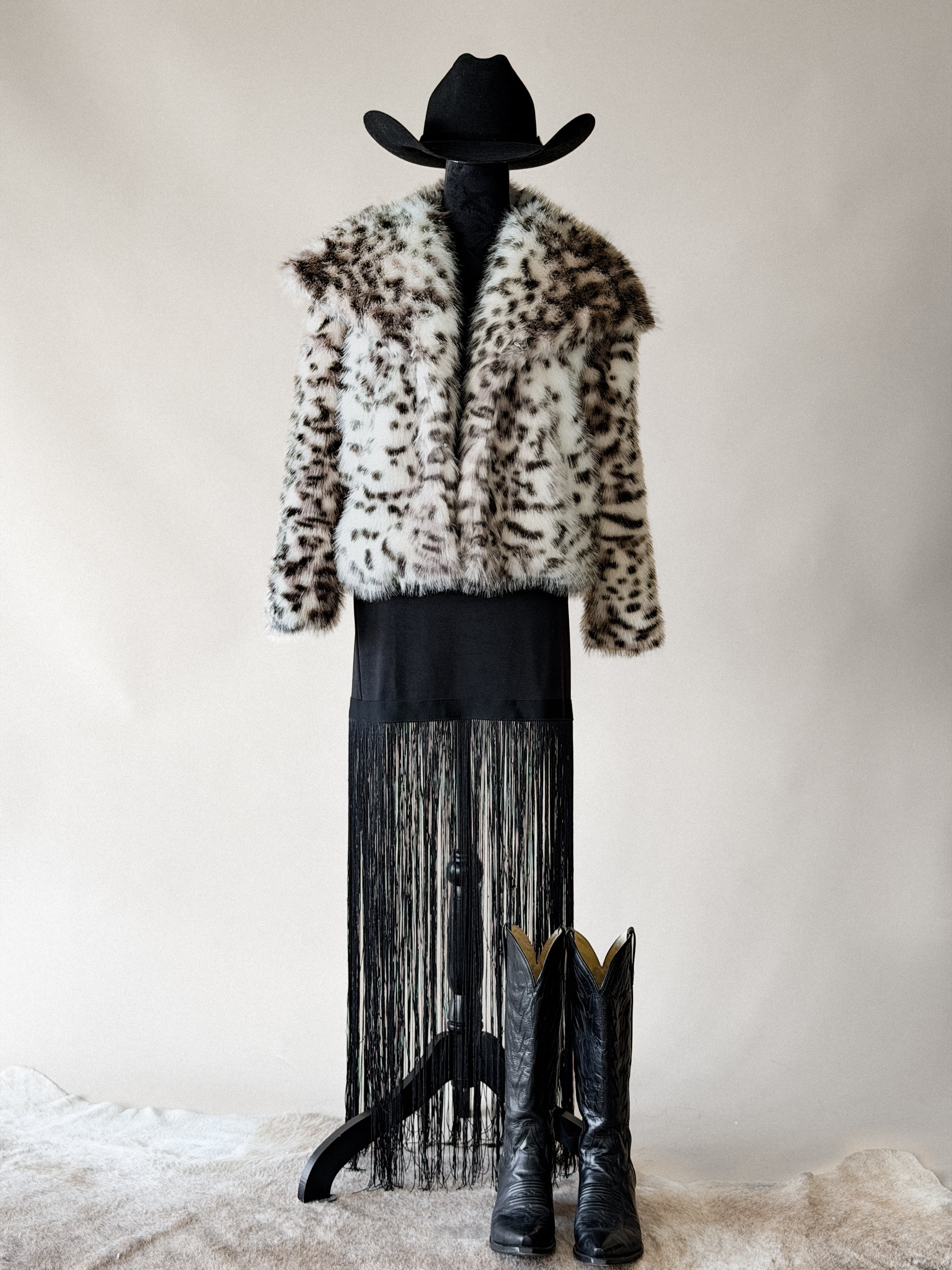 Western boho outfit with faux fur leopard print jacket, black fringe dress, cowboy boots, and hat