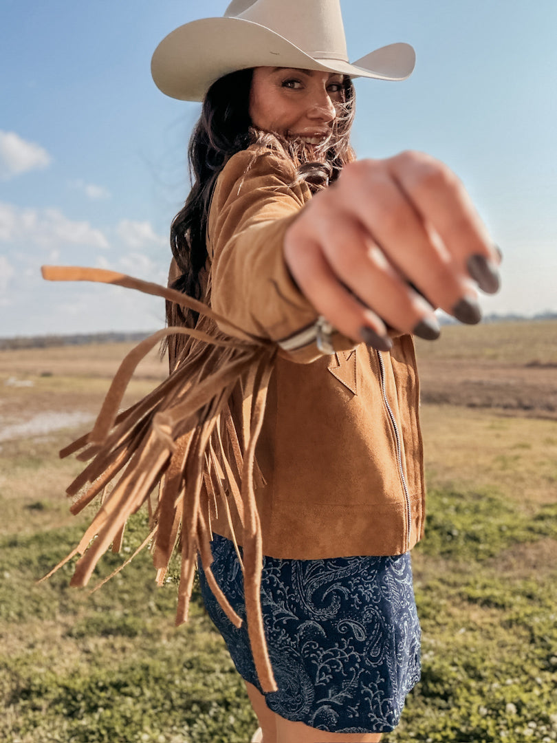 Woman in cowgirl hat and fringe suede jacket outdoors, wearing blue western boho skirt