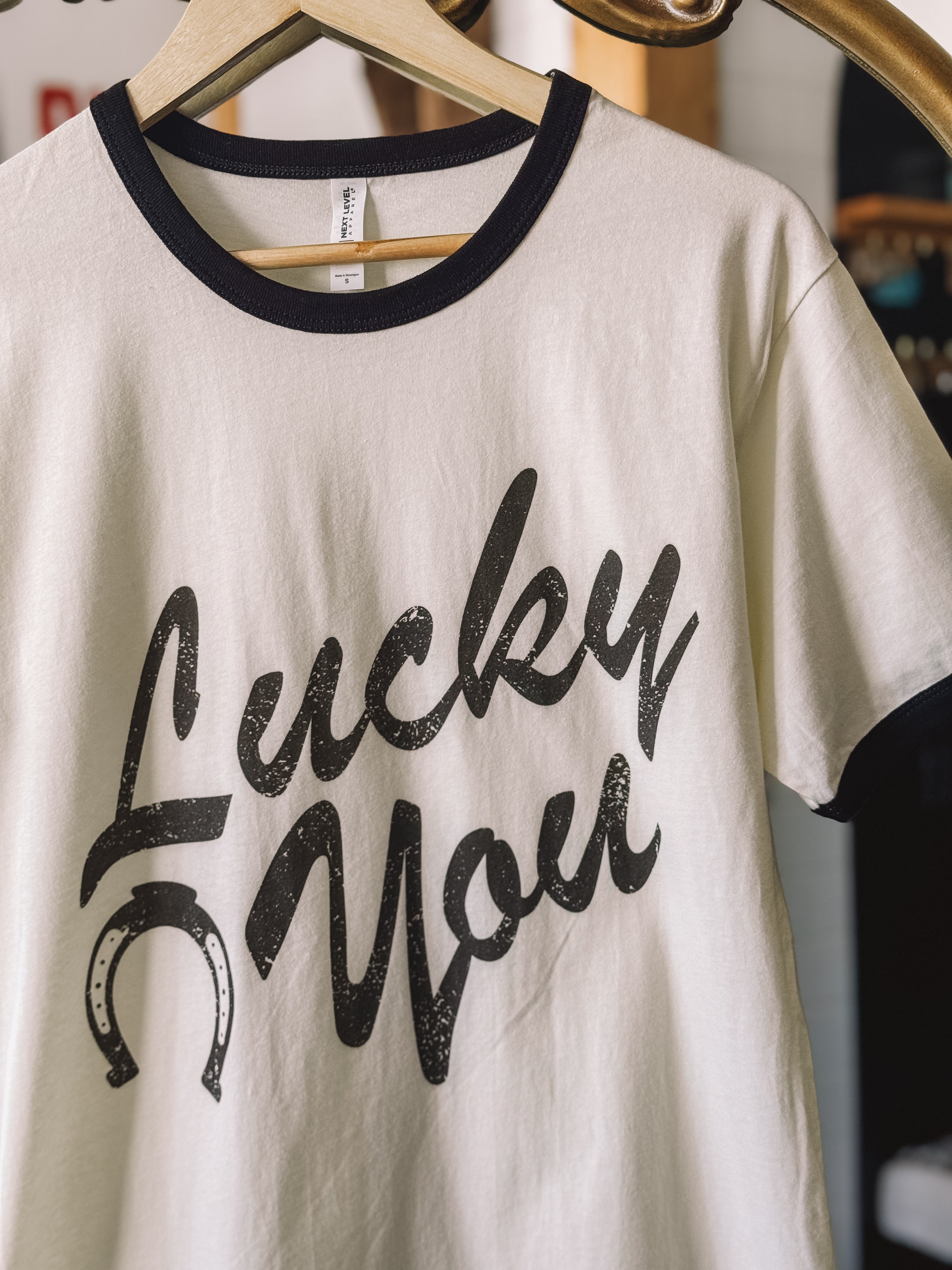 Western graphic tee with 'Lucky You' text and horseshoe, white with black trim, hanging on hanger
