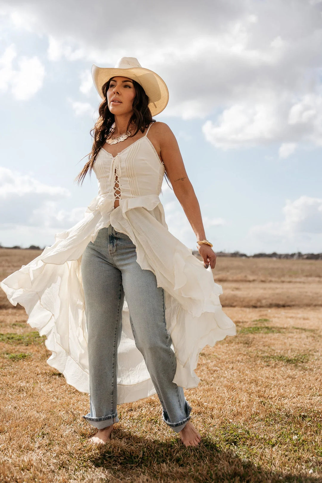 Woman in cowgirl hat, white boho top, and jeans posing barefoot in a western field