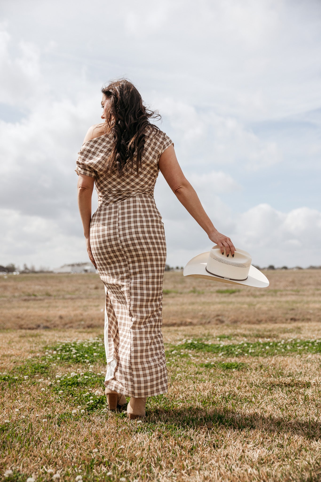 Woman in a brown plaid western dress holding a cowboy hat outdoors in a field