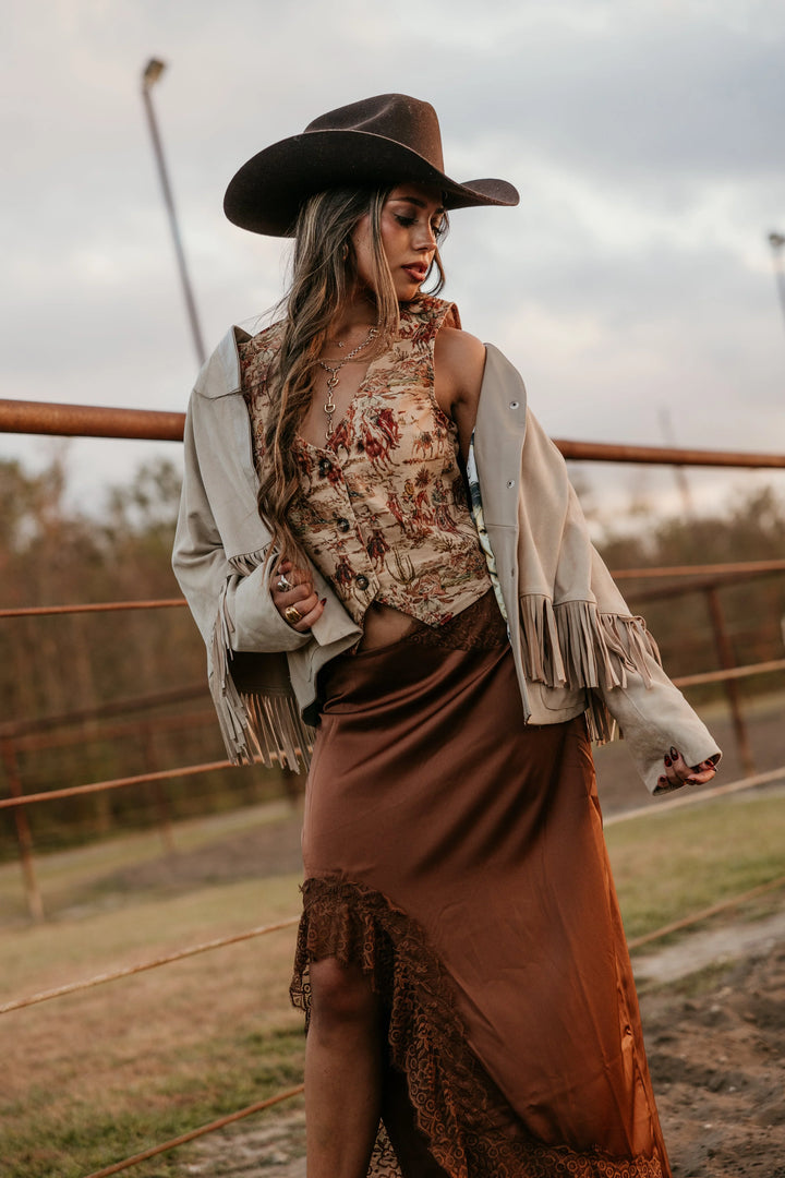 Woman in cowgirl hat, boho floral vest, fringe jacket, and satin skirt, western ranch setting