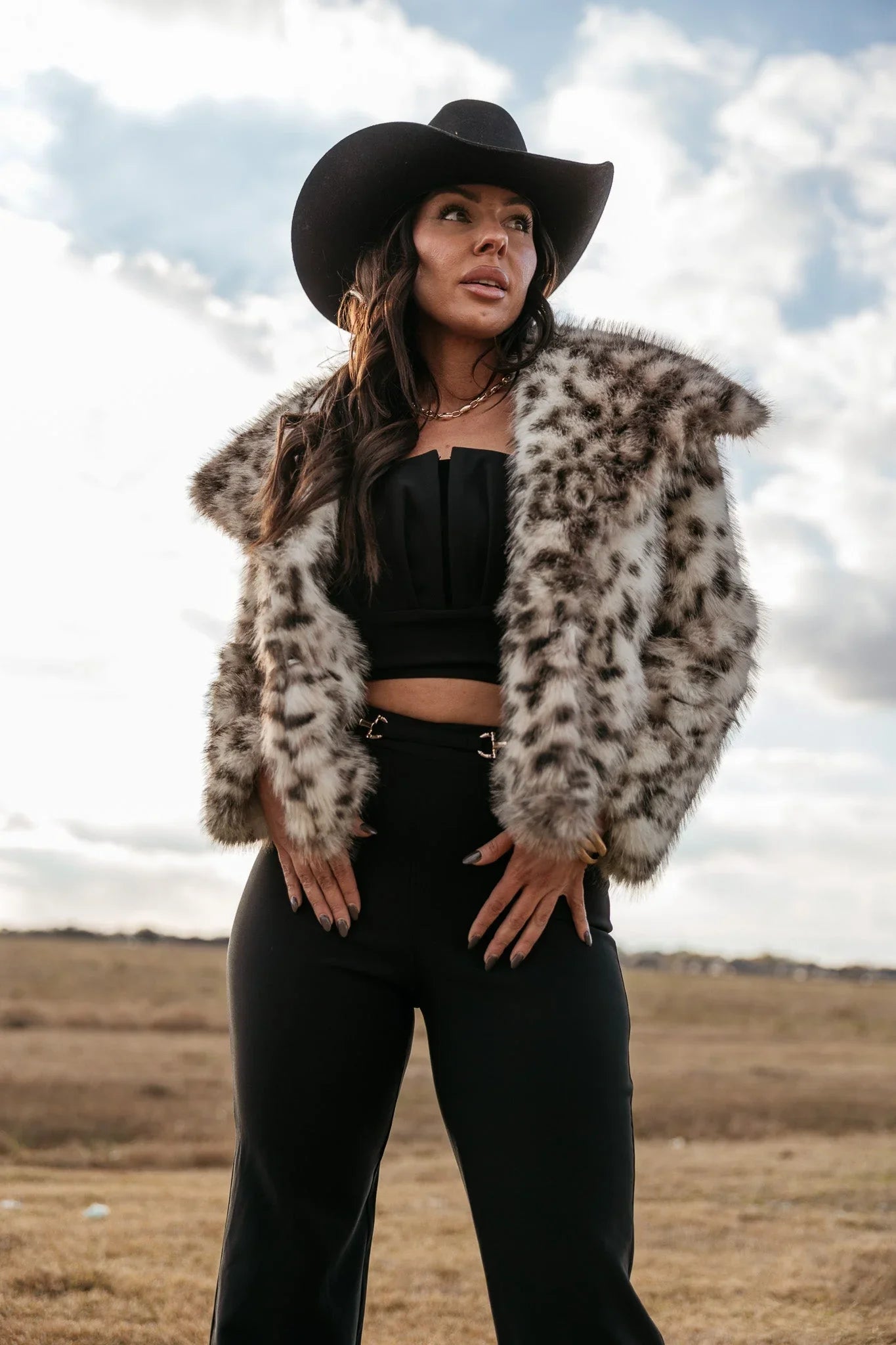 Woman in black cowgirl hat, faux fur animal print jacket, and western boho outfit outdoors