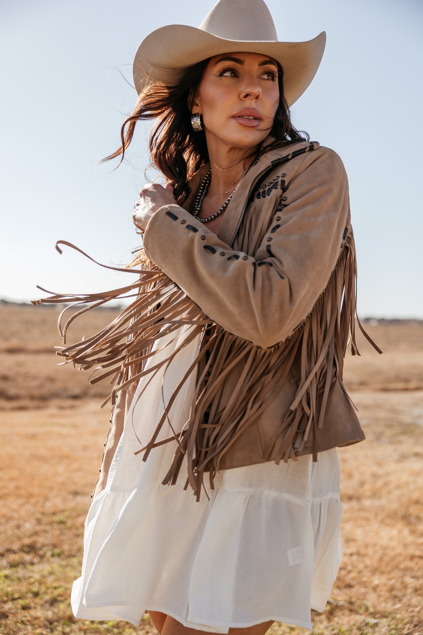 Woman in a fringed suede jacket, white western dress, and cowboy hat outdoors