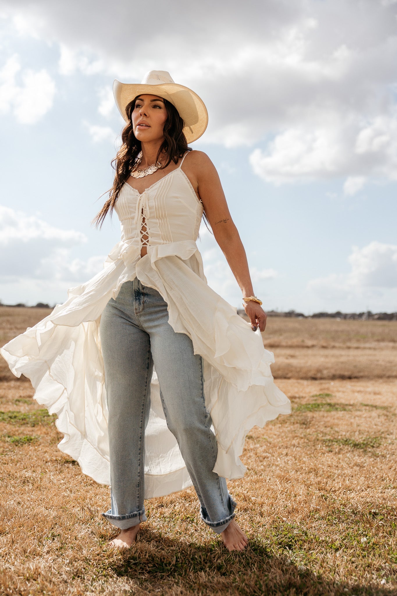 Woman in a white western lace-up top, cowboy hat, and blue jeans standing barefoot outdoors