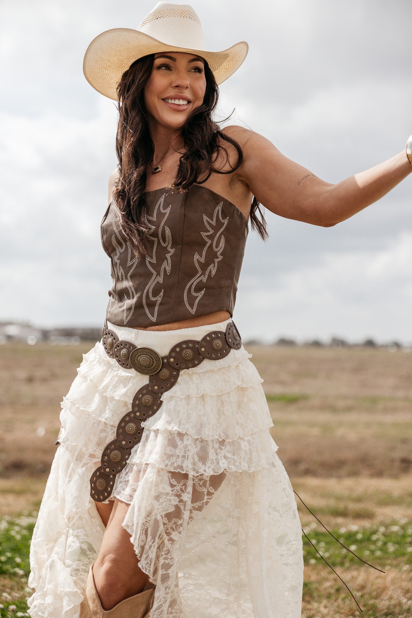 Woman in western boho outfit with cowboy hat, brown corset, lace skirt, and concho belt outdoors