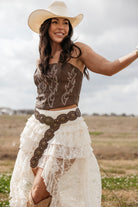 Woman in western boho outfit with cowboy hat, brown corset, lace skirt, and concho belt outdoors