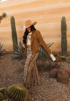 Woman in western boho outfit with wide-brim hat, fringe jacket, snakeskin pants, and turquoise jewelry in a desert setting with cacti.