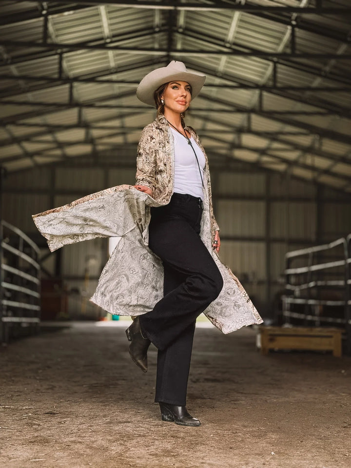 Woman in western boho outfit with cowboy hat, embroidered duster, black pants, and boots in barn