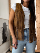 Woman wearing brown suede fringe vest, white t-shirt, light wash jeans, western jewelry