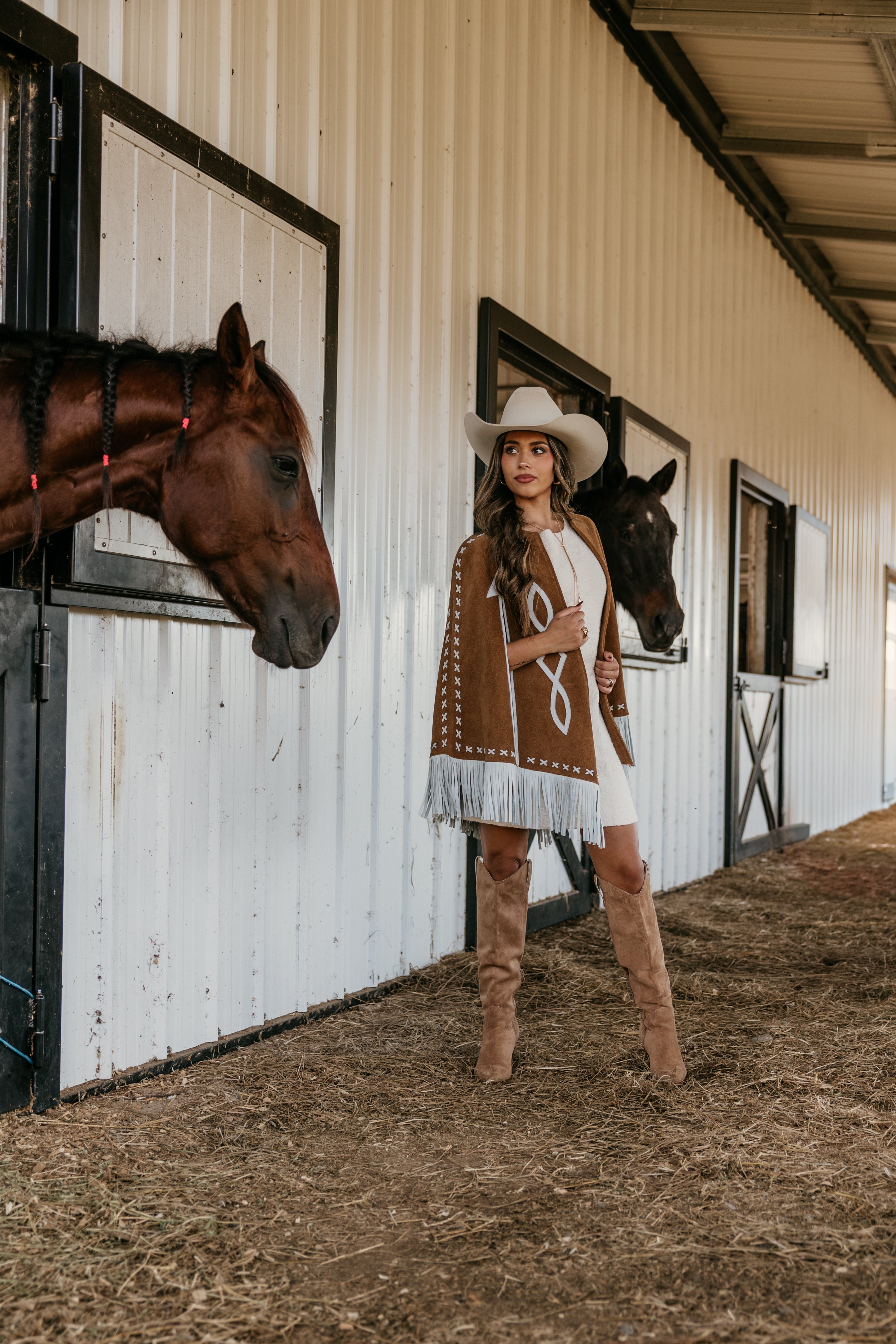 Woman in western boho outfit with cowboy hat and boots standing by horses in stable