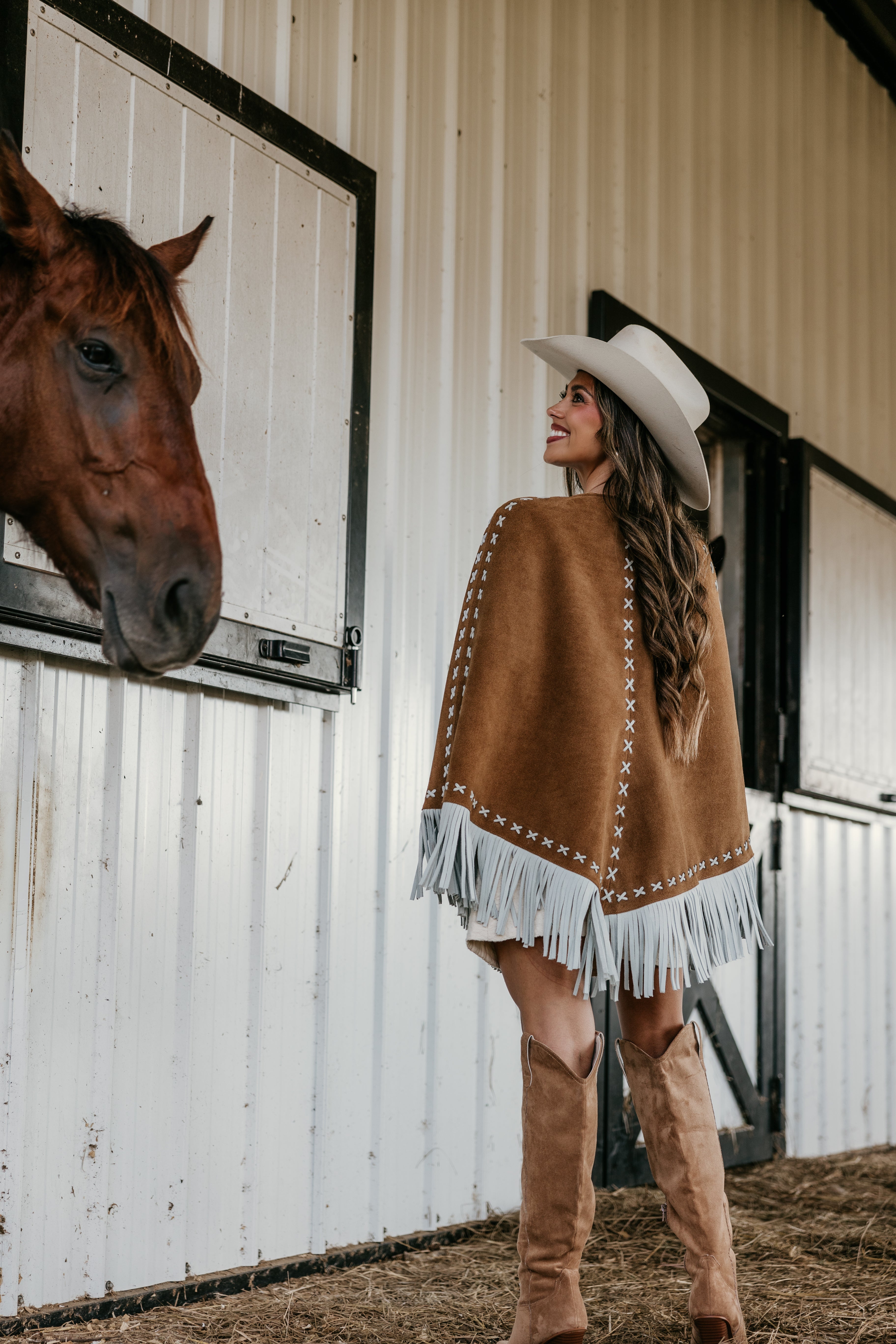 Woman in western fringe poncho, cowboy hat, and boots by horse barn, cowgirl outfit style