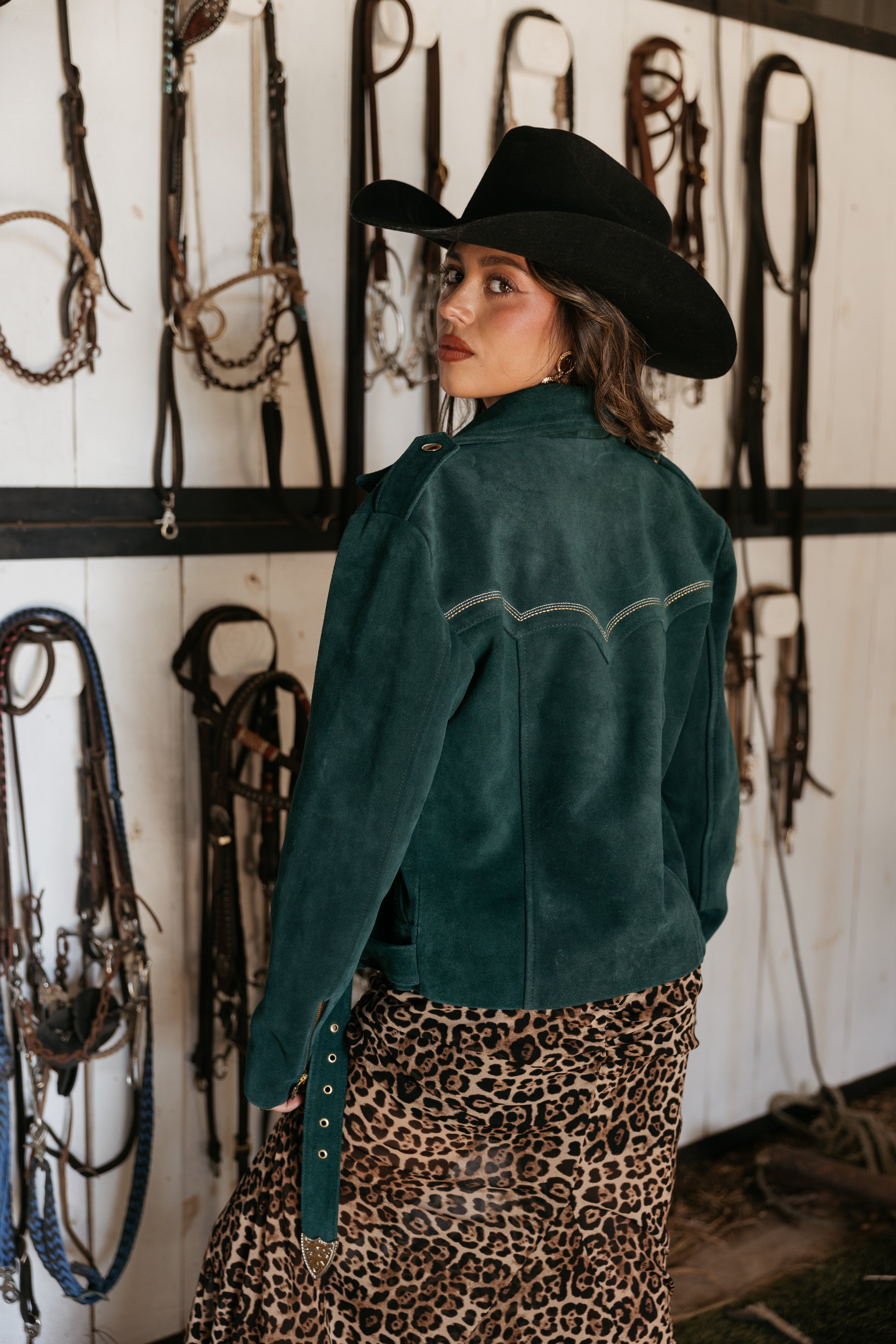Woman in a suede western jacket, black cowboy hat, and leopard print skirt in a tack room