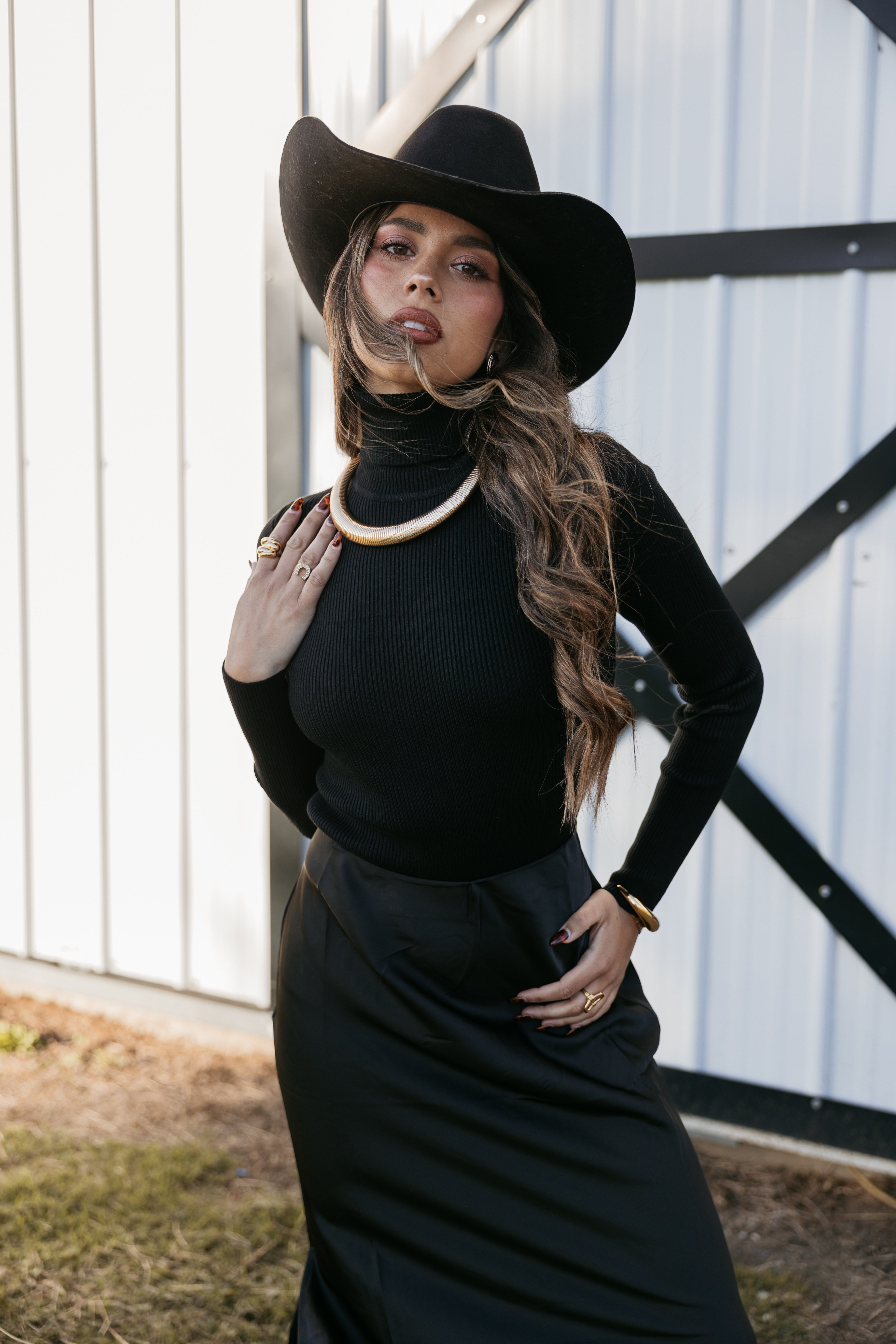 Woman in black turtleneck, cowboy hat, and western jewelry posing by barn door
