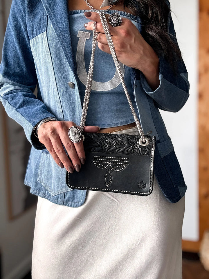Person holding a black cowhide handbag with intricate designs, wearing a blue denim jacket and white pants.