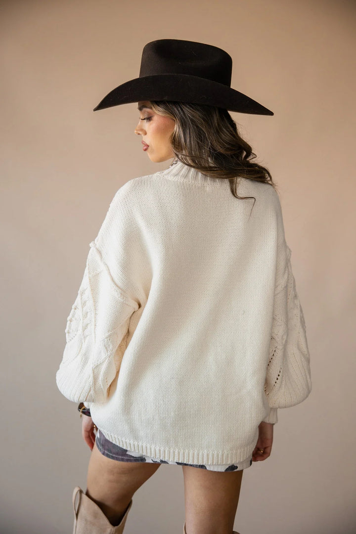 Woman in a cream knit sweater, brown cowboy hat, western boots, and patterned skirt