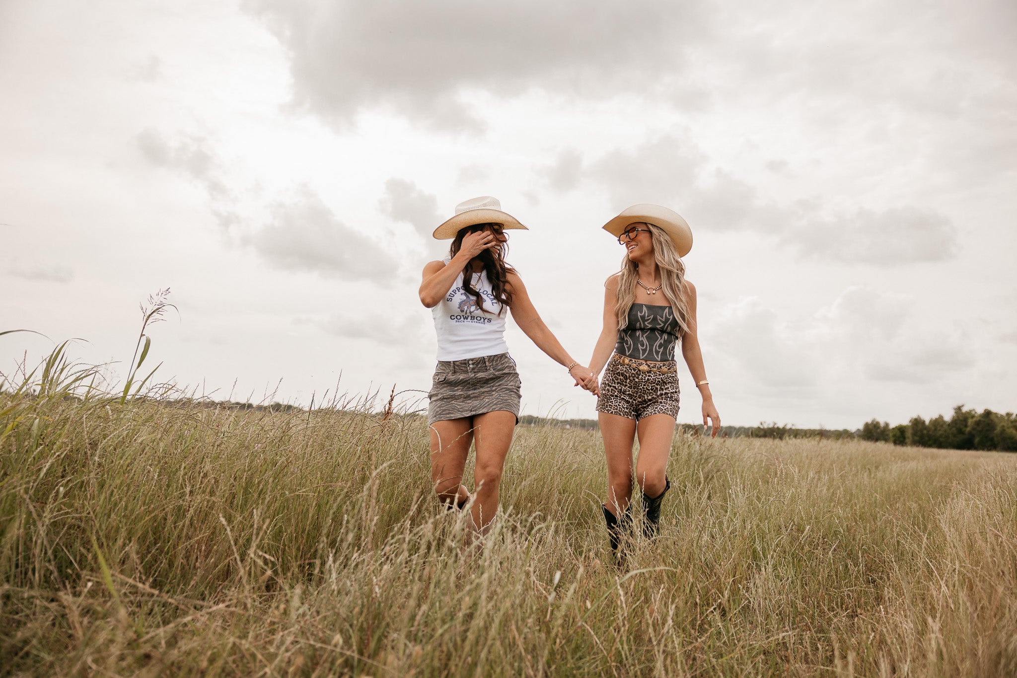 Two women walking hand in hand through a field with a cloudy sky.