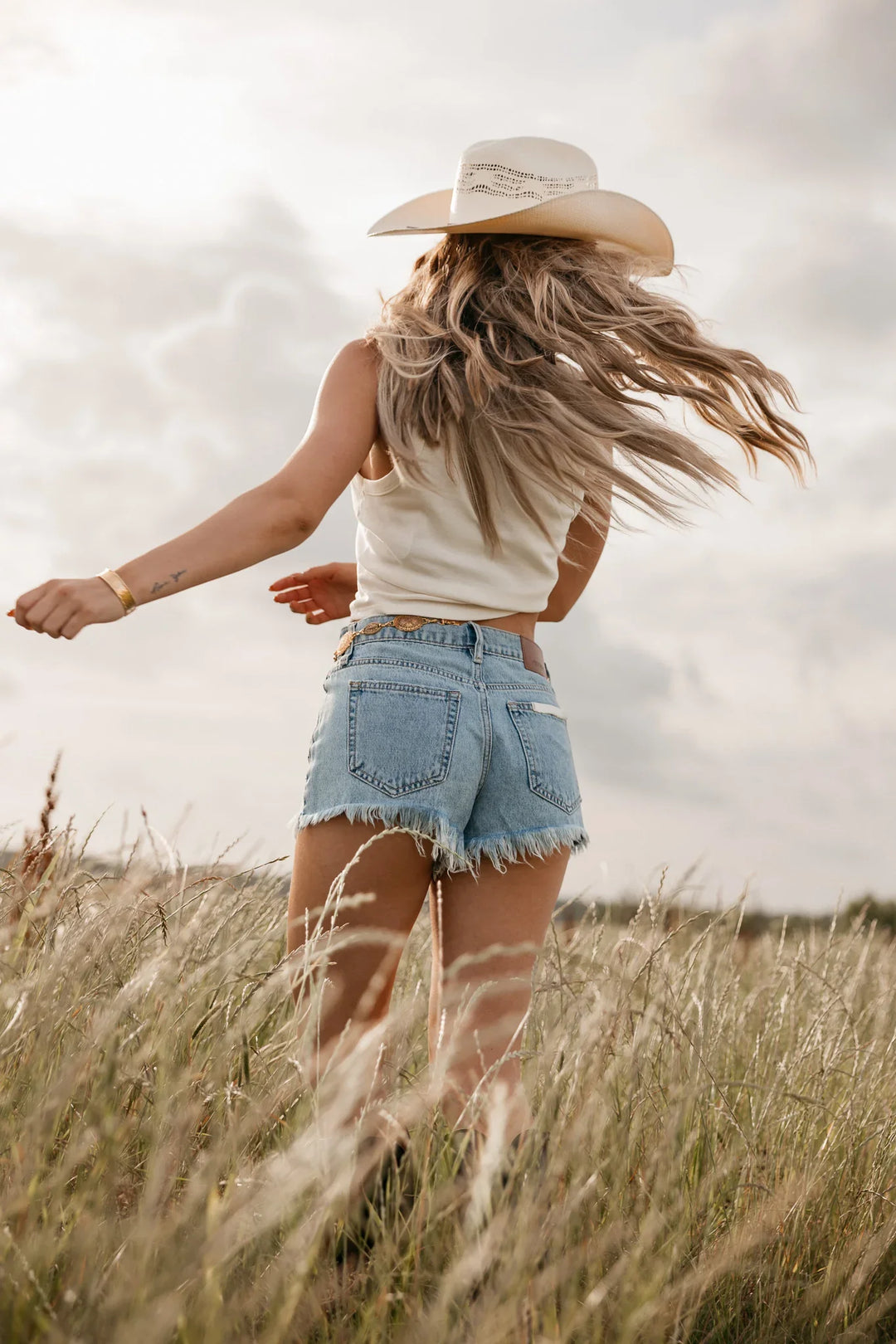 Woman in a cowboy hat and denim shorts standing in a field with long hair blowing in the wind.