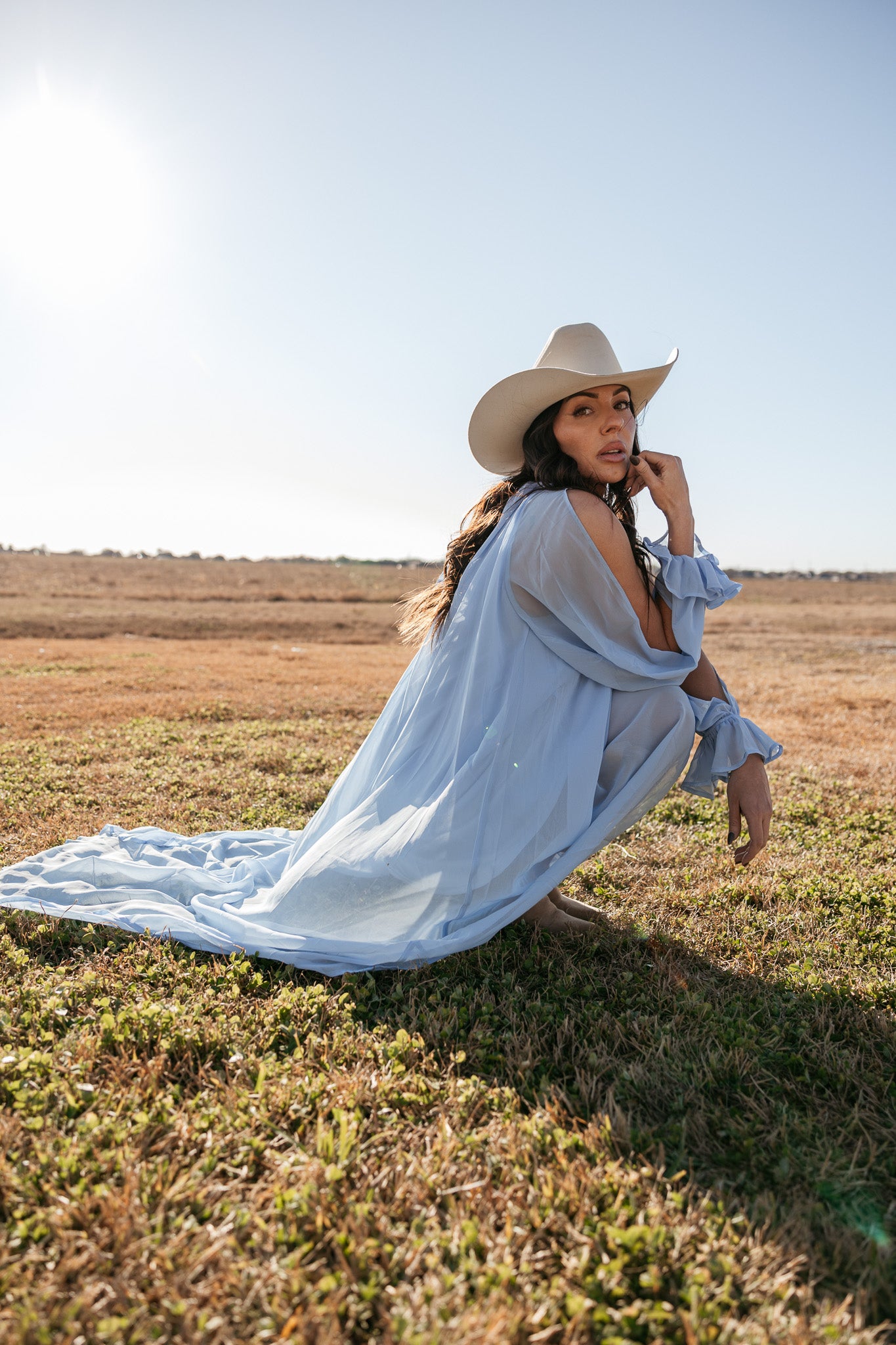 Woman in a long light blue dress and cowboy hat sitting in a field.