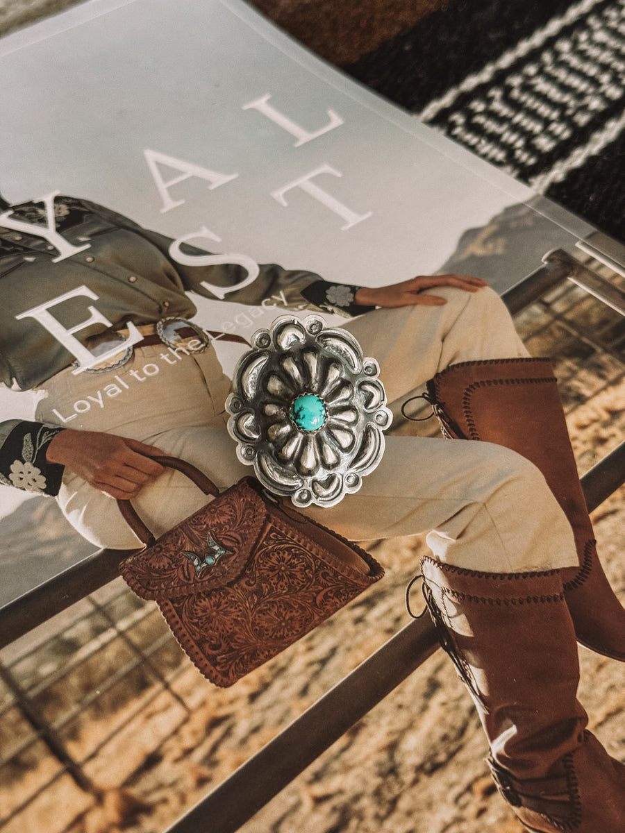 A Navajo Concho Ring with a turquoise stone at its center, displayed on a person's finger.